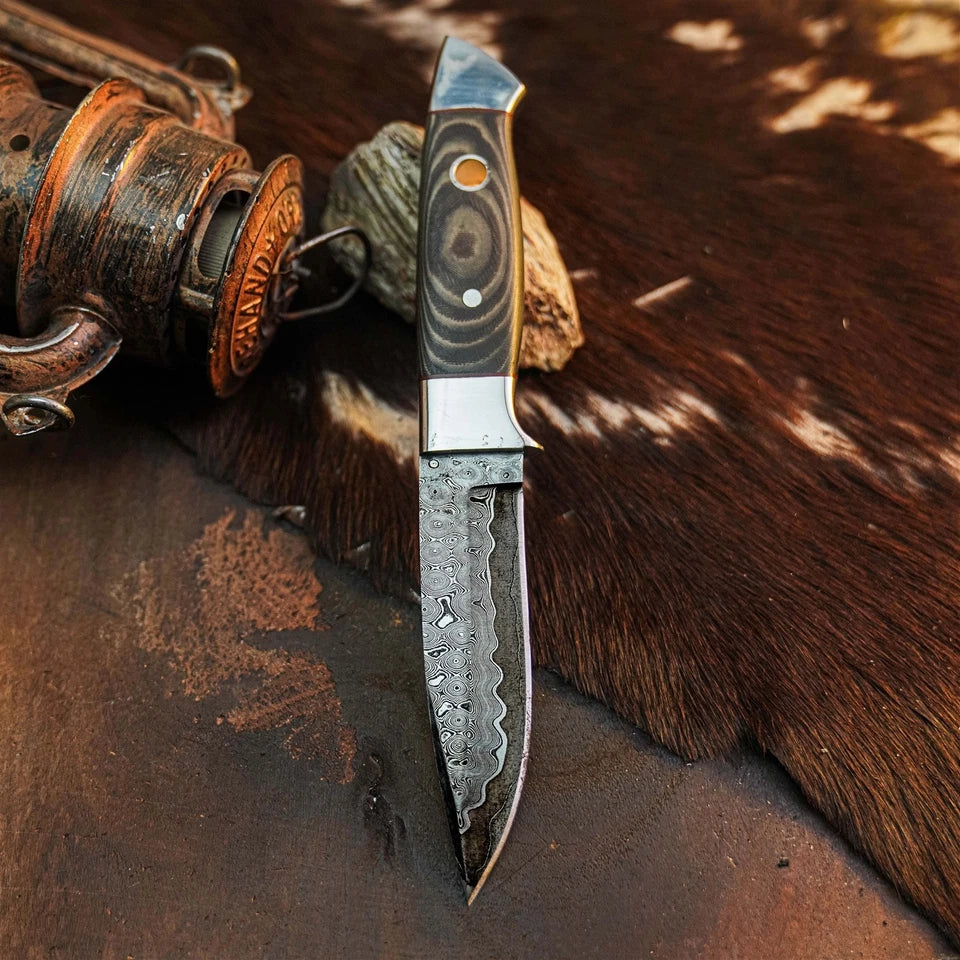 A custom handmade fixed-blade knife with a textured, hammered Damascus steel blade and a patterned black and silver handle, displayed on a rustic brown leather and cowhide surface next to a vintage wooden object.