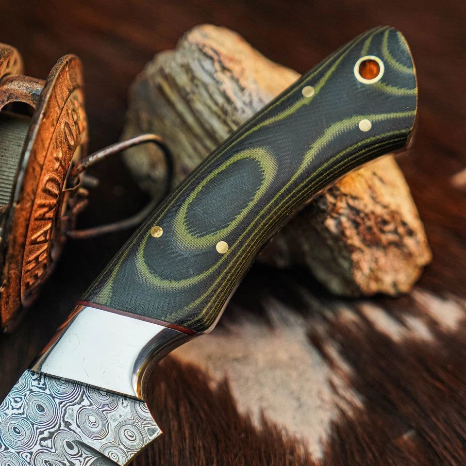 A close-up of a knife handle with a dark green and black wood grain pattern, brass pins, and a silver bolster, resting on rocks over a brown hide background.