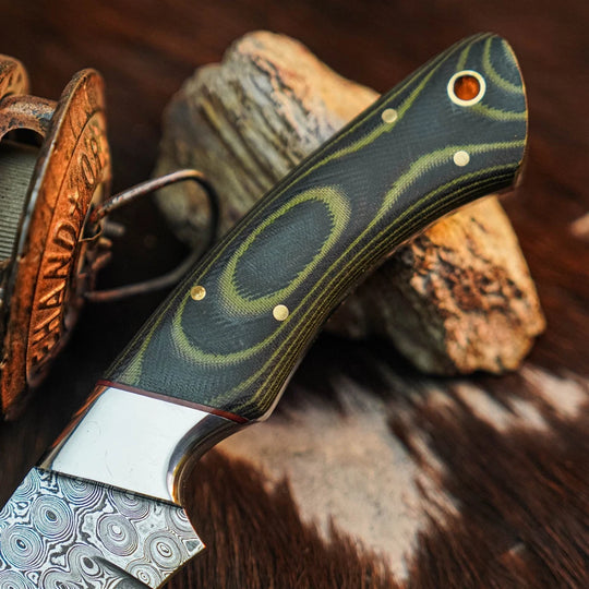 A close-up of a knife handle with a dark green and black wood grain pattern, brass pins, and a silver bolster, resting on rocks over a brown hide background.