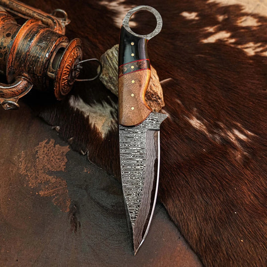 A fixed-blade knife with a patterned Damascus-style steel blade, a brown and black handle, and a ring pommel, photographed upright on a brown-and-white animal hide with rustic metal props nearby.