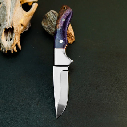 A fixed-blade knife with a polished silver steel blade and a purple-toned wood handle, displayed on a dark textured surface next to animal skull and wood props.