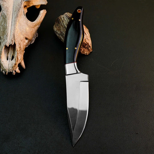 A fixed-blade knife with a polished silver blade and a black handle accented with gold pins, displayed on a dark textured surface next to a bone skull prop.