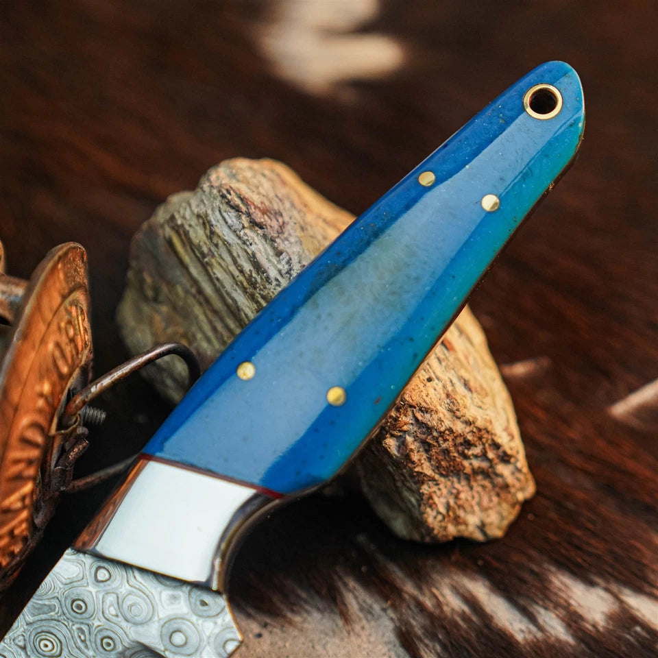 A close-up of a knife with a glossy blue handle and brass pins, resting on rustic wood and stone against a dark brown textured background.