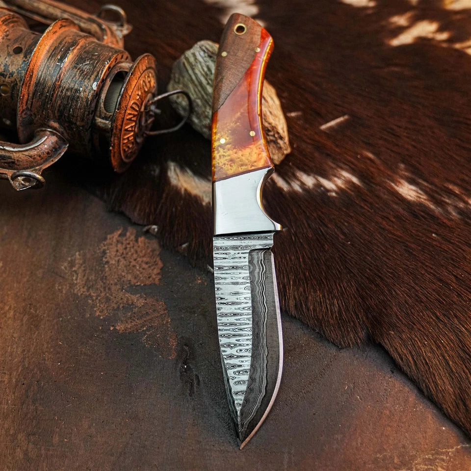 A fixed-blade knife with a patterned Damascus-style steel blade and a reddish-brown wood handle, displayed on a dark brown textured surface with a cowhide rug and a rustic metal container nearby.