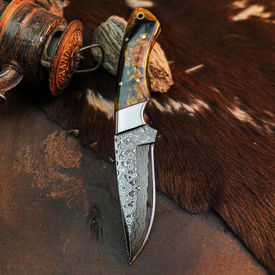 A custom fixed-blade knife with a patterned Damascus steel blade and a carved handle made of brown and gold-toned wood, displayed on a dark textured surface next to a leather and wood mallet.