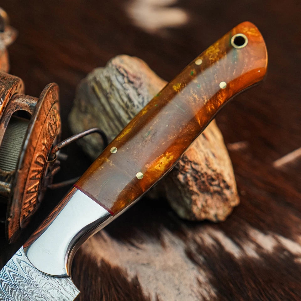 A close-up of a custom knife with a polished Damascus steel blade and a marbled amber-brown handle, resting on rustic wood and leather.