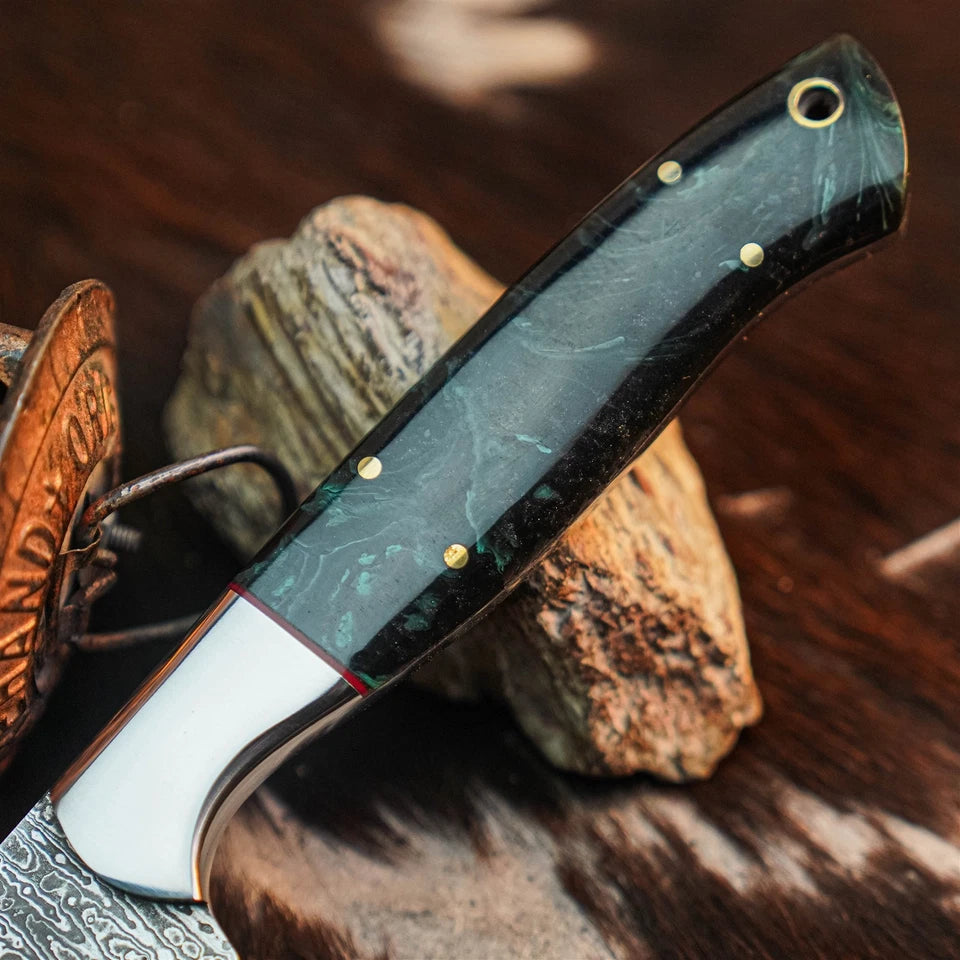 A close-up of a folding pocket knife with a dark green and black marbled handle, brass pins, and a white bolster, resting on natural stones over a dark textured surface.