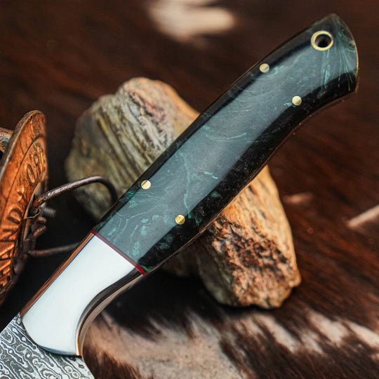 A close-up of a folding pocket knife with a dark green and black marbled handle, brass pins, and a white bolster, resting on natural stones over a dark textured surface.