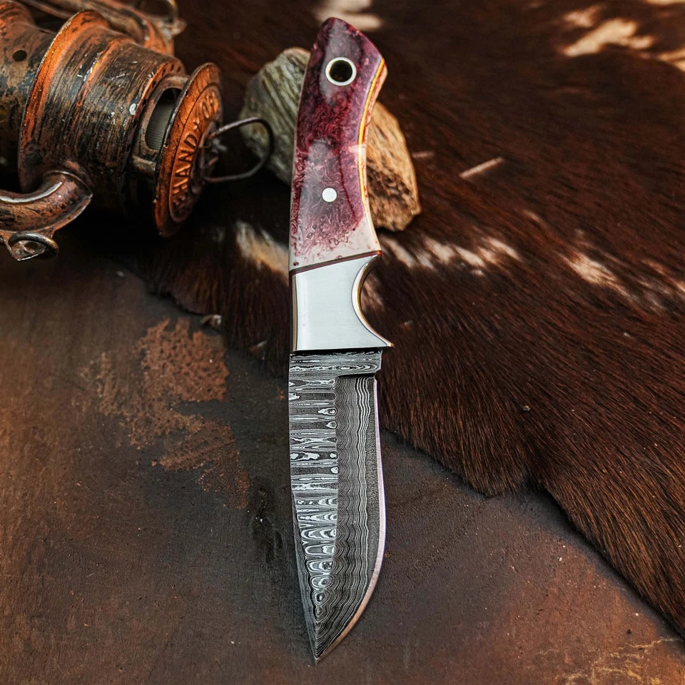 A custom fixed-blade knife with a Damascus steel blade and a reddish-brown wood handle, displayed on a rustic brown leather and hide background.