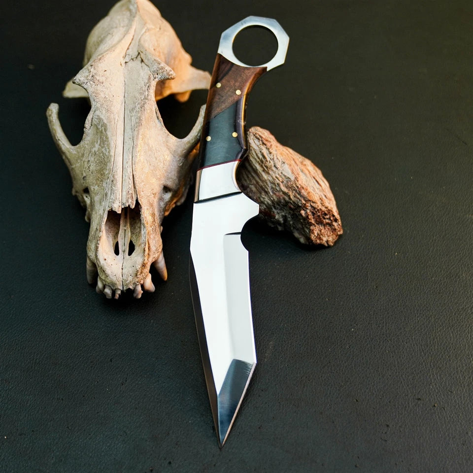 A fixed-blade knife with a polished silver blade, dark handle, and wood inlay, displayed diagonally next to a deer skull and driftwood on a dark textured surface.