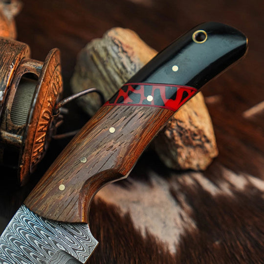 A close-up of a Damascus steel knife with a richly carved wooden handle, featuring a black inlay, red patterned accent, and brass pins, resting on rocks and fur against a rustic wooden background.