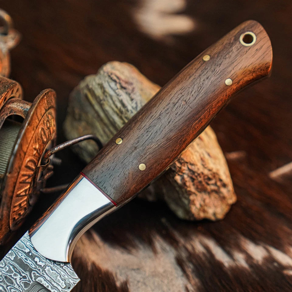 A close-up of a fixed-blade knife with a dark brown wooden handle and patterned steel blade, resting on rustic wood and leather.
