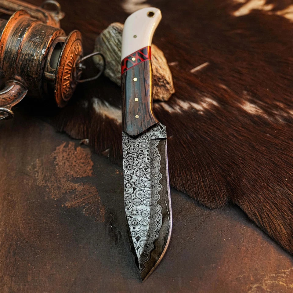 A fixed-blade knife with a patterned Damascus steel blade, a dark wood handle, and a white handle end, displayed on a rustic brown surface with a cowhide and a metal container nearby.