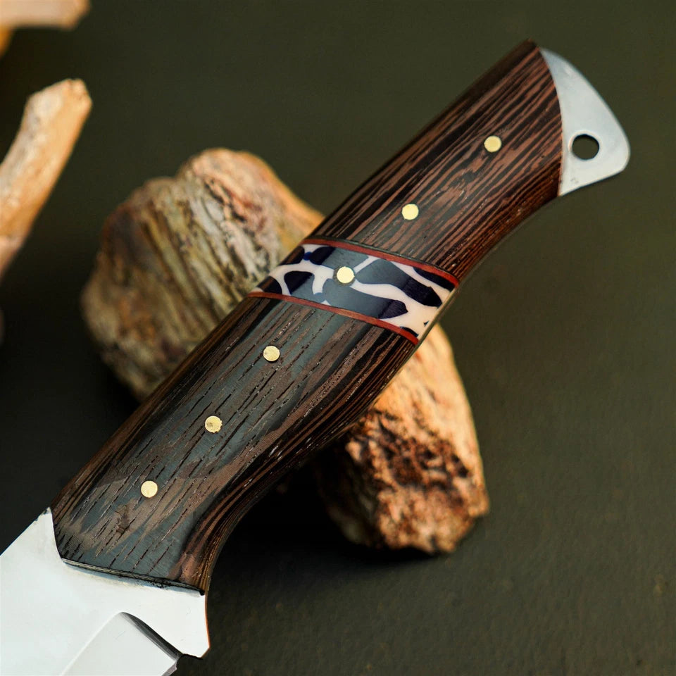 A close-up of a handcrafted knife with a dark wood handle, featuring brass pins, a black and white patterned inlay band, and a silver-colored metal bolster, resting on rustic wood pieces against a dark background.