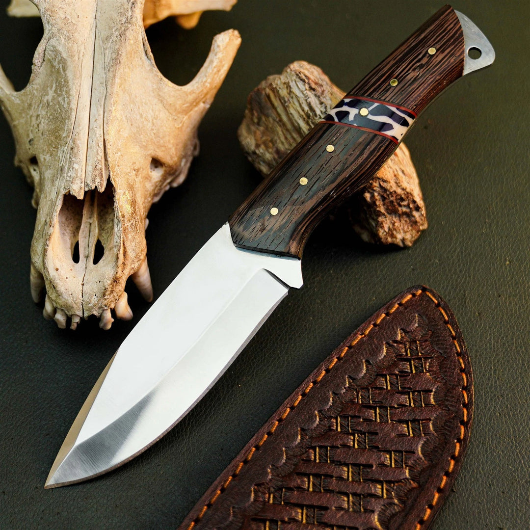 A fixed-blade knife with a polished silver blade and a dark brown wood-grain handle with brass pins and a red accent band, displayed diagonally next to its brown leather sheath on a dark textured surface, with a bone skull and rock props in the background.
