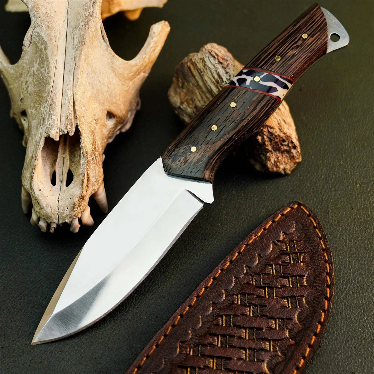 A fixed-blade knife with a polished silver blade and a dark brown wood-grain handle with brass pins and a red accent band, displayed diagonally next to its brown leather sheath on a dark textured surface, with a bone skull and rock props in the background.