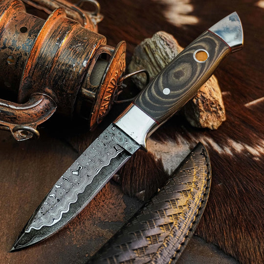 A custom knife with a patterned Damascus steel blade and a dark wood handle with a silver bolster, displayed diagonally on a rustic background with a leather sheath, a vintage fishing reel, and rocks.
