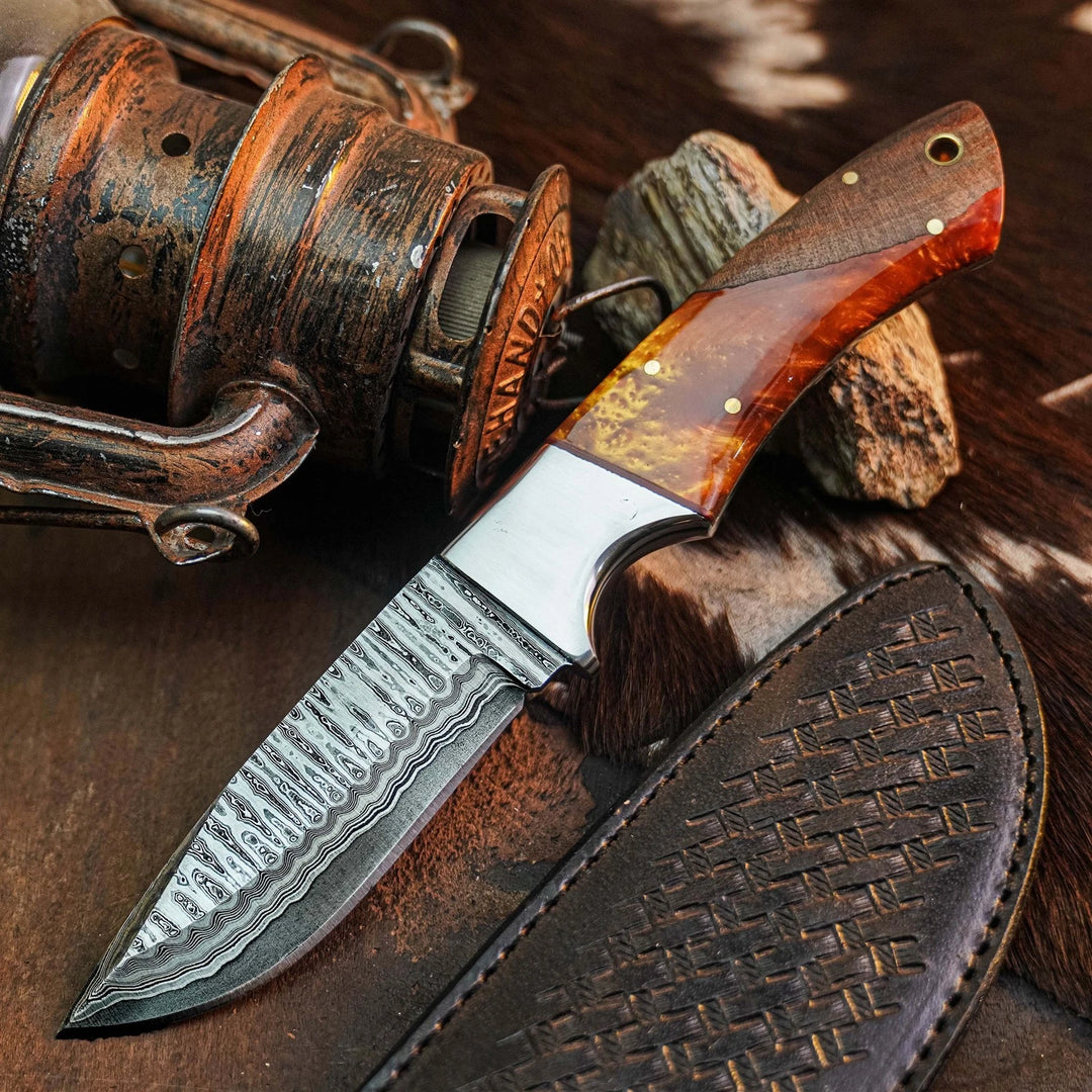 A custom fixed-blade knife with a patterned Damascus steel blade, a polished silver bolster, and a reddish-brown wood handle with brass pins, displayed next to its dark brown leather sheath on a rustic background with a vintage fishing reel.