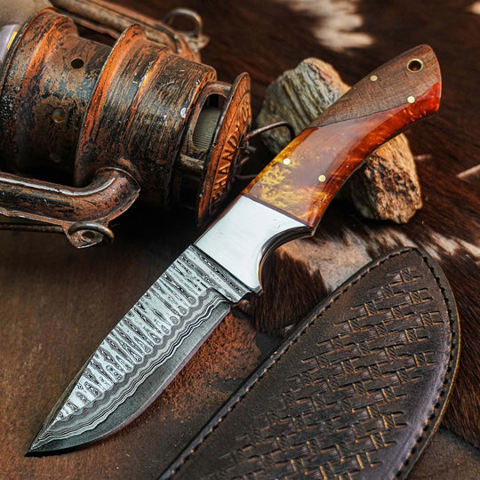 A custom fixed-blade knife with a patterned Damascus steel blade, a polished silver bolster, and a reddish-brown wood handle with brass pins, displayed next to its dark brown leather sheath on a rustic background with a vintage fishing reel.