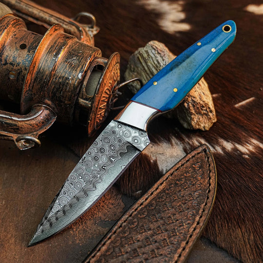 A fixed-blade knife with a patterned Damascus steel blade and a bright blue handle with brass pins, displayed on a rustic brown fur surface with a leather sheath and vintage metal object nearby.