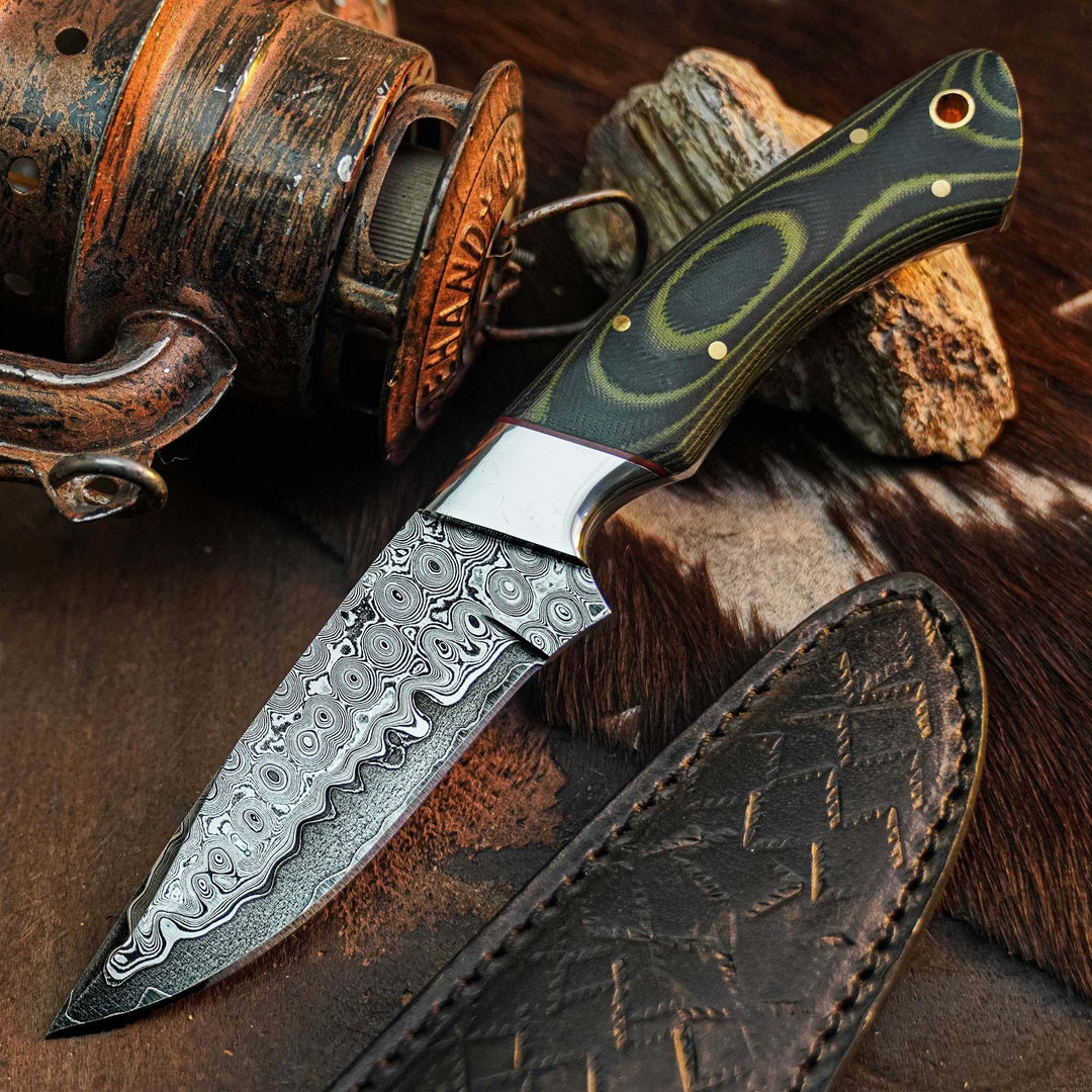 A fixed-blade knife with a patterned Damascus steel blade, a dark green and black handle with gold accents, and a brown leather sheath, styled on a rustic background with wood and a fishing reel.