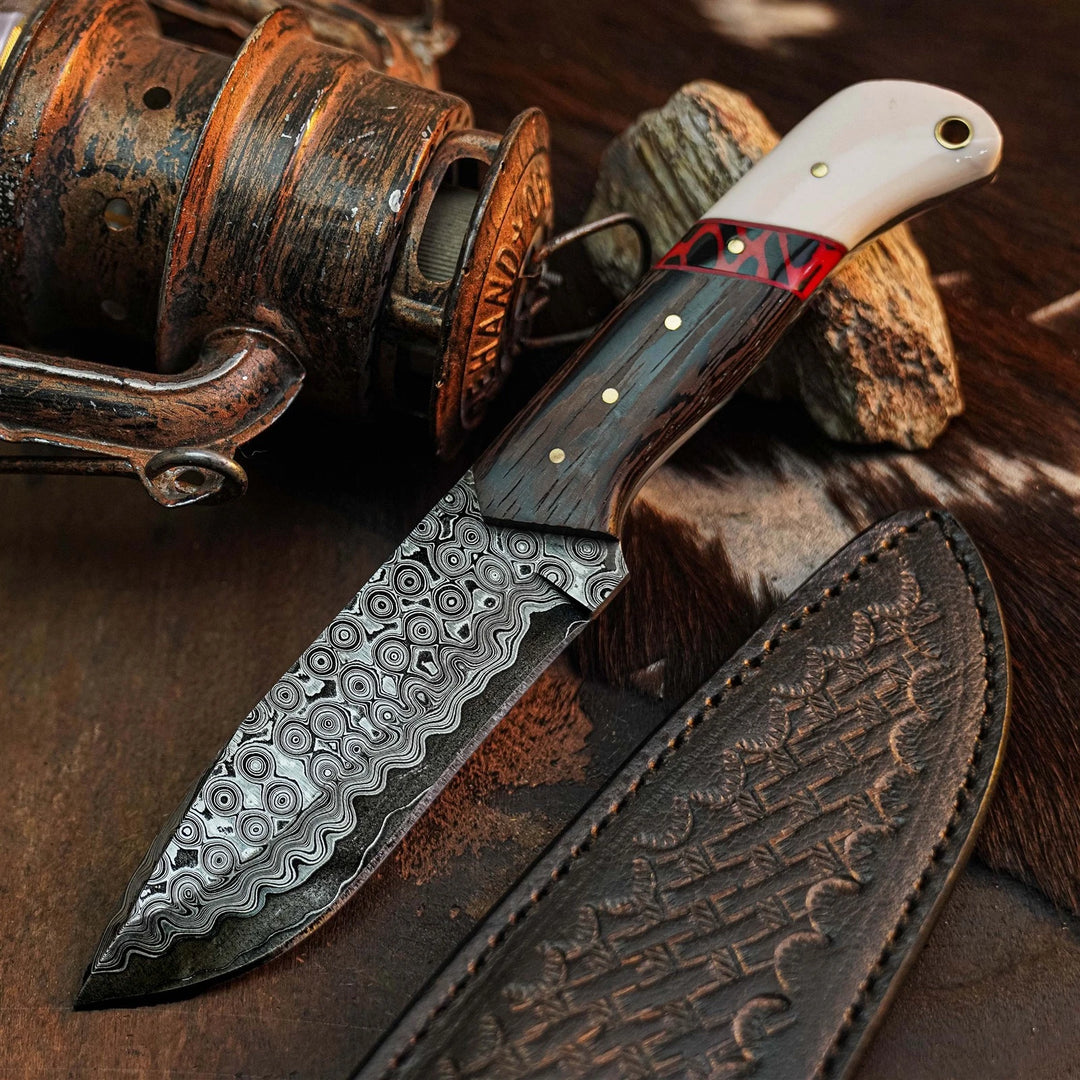 A fixed-blade knife with a patterned Damascus-style steel blade, a dark wood handle with a red inlay and brass pins, and a white horn or bone section near the bolster. A matching embossed brown leather sheath lies beside it on a rustic brown surface, with a fishing reel and rock props in the background.