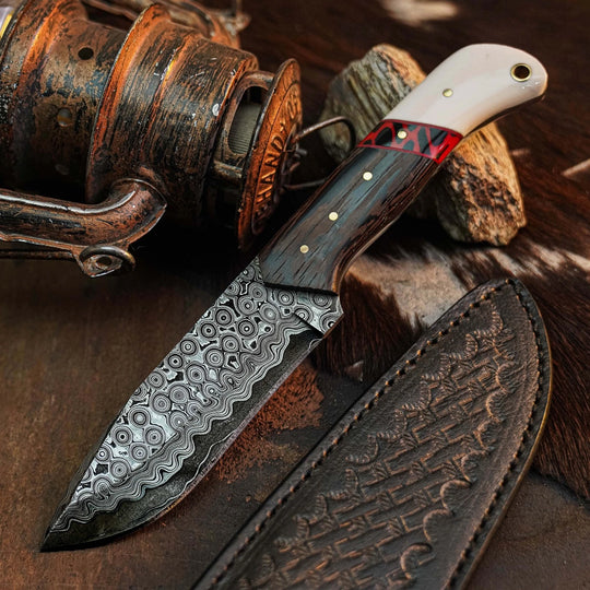 A fixed-blade knife with a patterned Damascus-style steel blade, a dark wood handle with a red inlay and brass pins, and a white horn or bone section near the bolster. A matching embossed brown leather sheath lies beside it on a rustic brown surface, with a fishing reel and rock props in the background.