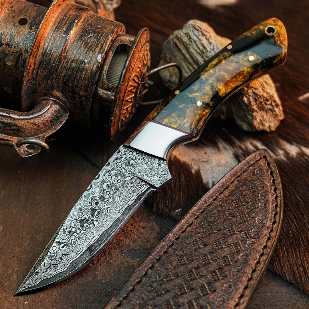 A fixed-blade knife with a patterned Damascus-style steel blade and a marbled amber-brown handle, shown with a brown leather sheath on a rustic wood and fur surface. A vintage fishing reel appears in the background as a styled prop.