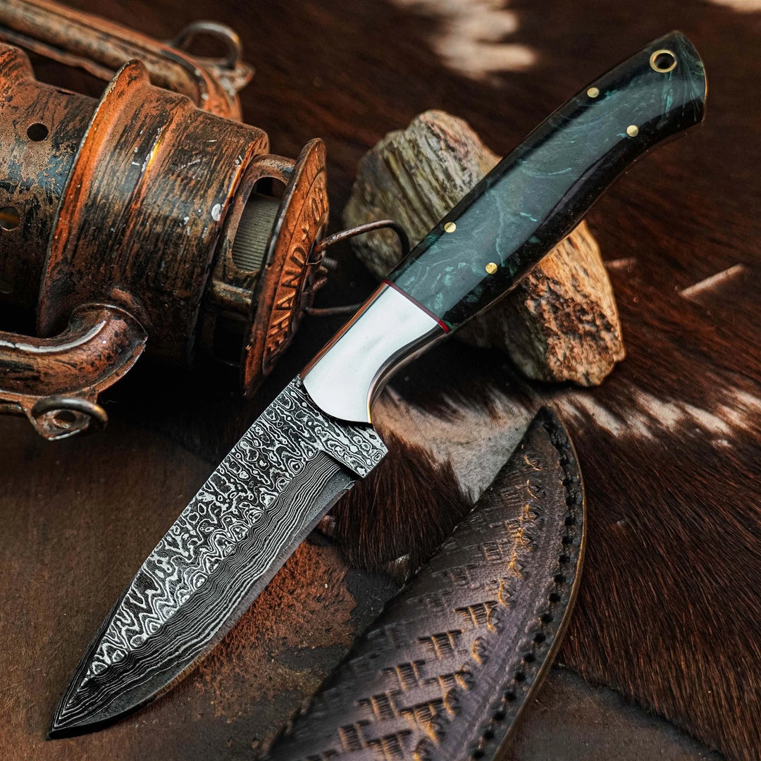 A fixed-blade knife with a patterned Damascus-style steel blade, a polished silver bolster, and a dark green marbled handle, displayed diagonally on a rustic brown leather surface with a vintage fishing reel and rock props.