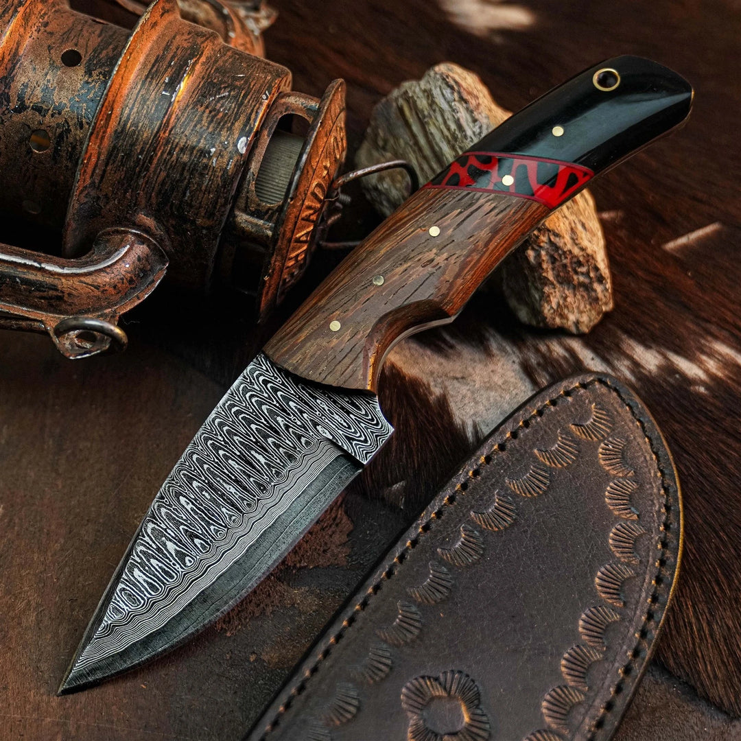 A Damascus steel fixed-blade knife with a patterned steel blade, a contoured wooden handle with a red and black inlay, and brass pins, shown next to its brown leather sheath on a rustic background with a vintage fishing reel.