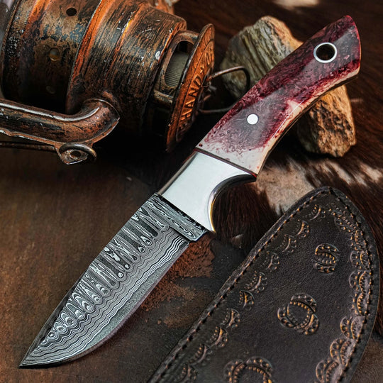 A fixed-blade knife with a patterned Damascus-style steel blade, a polished silver bolster, and a reddish-brown marbled handle, shown with a dark brown tooled leather sheath on a rustic brown surface. A vintage copper reel and rock are partially visible in the background.
