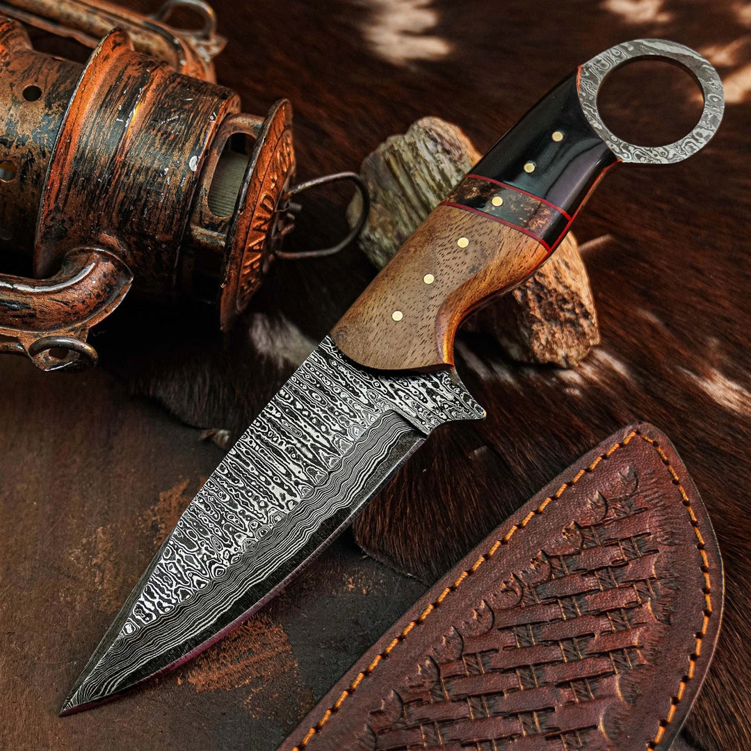 A fixed-blade knife with a patterned Damascus steel blade, a brown wood handle, and a black handle end, displayed diagonally on a rustic brown fur surface next to a leather sheath and a vintage fishing reel.