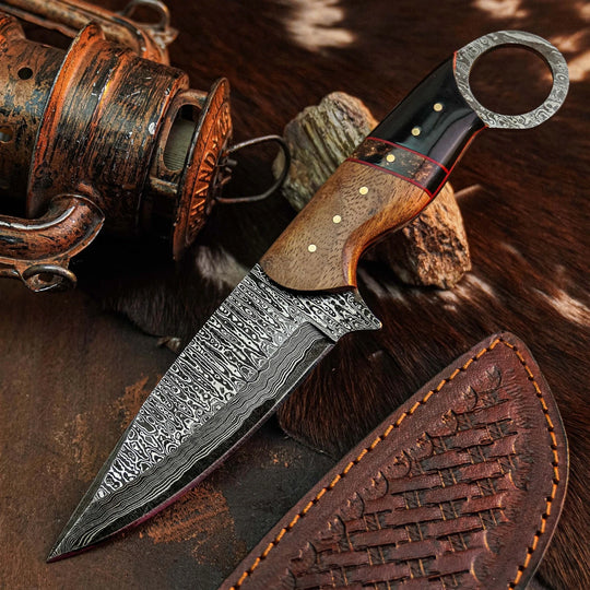 A fixed-blade knife with a patterned Damascus steel blade, a brown wood handle, and a black handle end, displayed diagonally on a rustic brown fur surface next to a leather sheath and a vintage fishing reel.