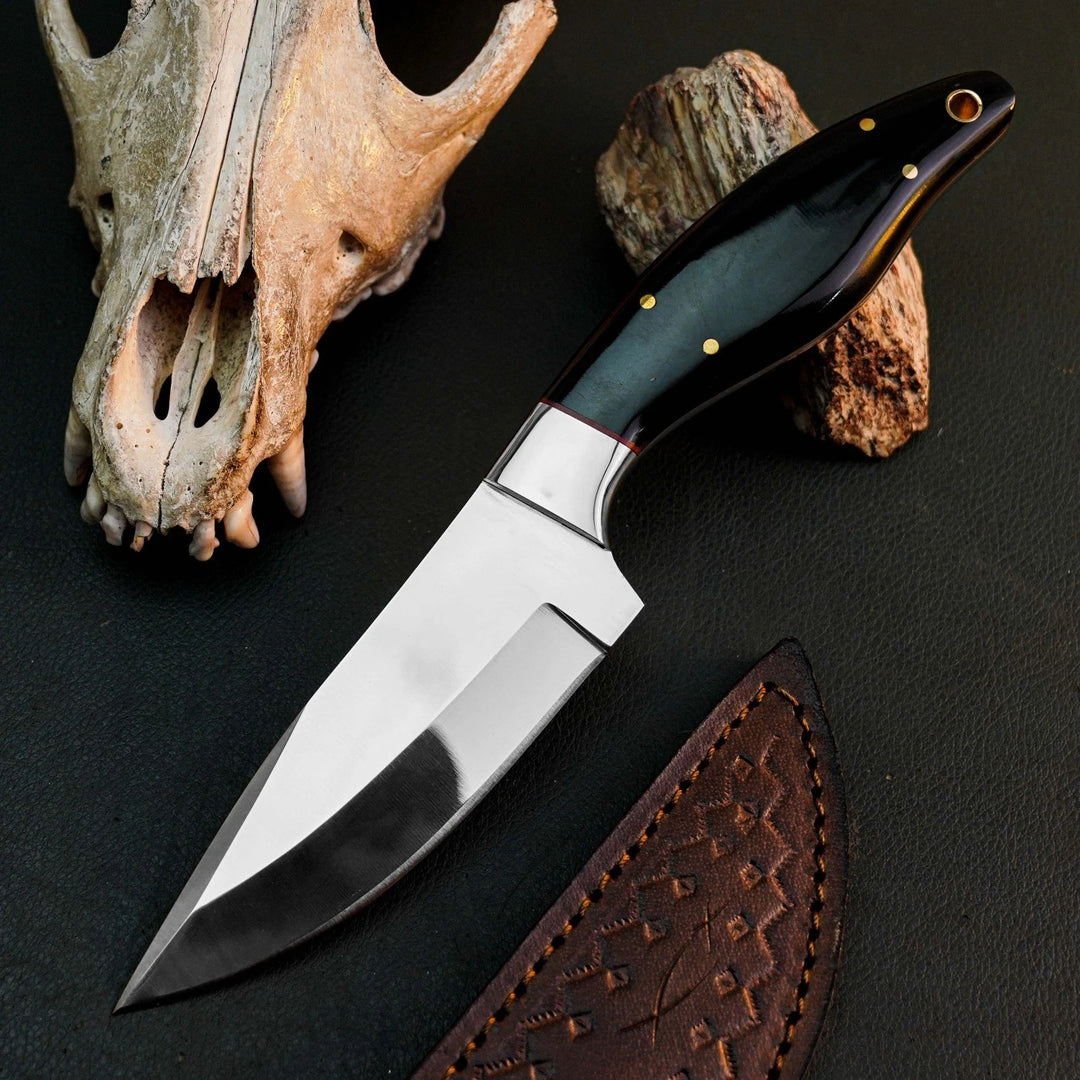 A fixed-blade knife with a polished silver blade and a dark green handle with brass pins, displayed diagonally on a dark textured surface next to a brown leather sheath, a skull, and a rock.