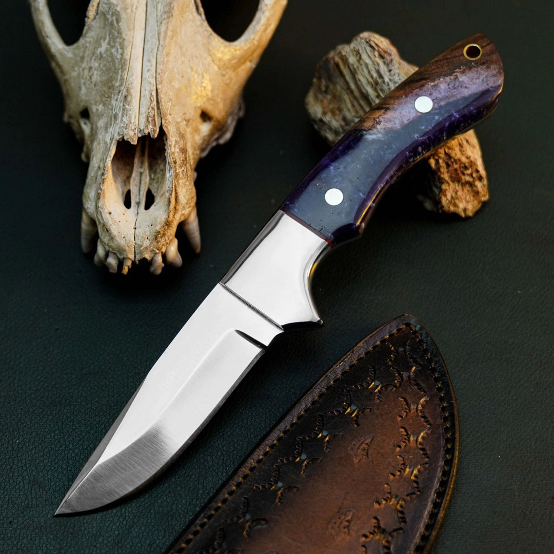 A fixed-blade knife with a satin-finished silver blade and a dark purple marbled handle, displayed diagonally next to its brown leather sheath. The background is a dark textured surface decorated with a weathered animal skull and a piece of driftwood.