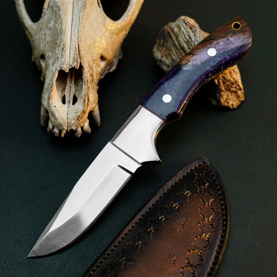 A fixed-blade knife with a satin-finished silver blade and a dark purple marbled handle, displayed diagonally next to its brown leather sheath. The background is a dark textured surface decorated with a weathered animal skull and a piece of driftwood.