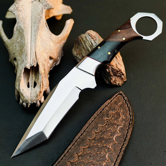 A fixed-blade knife with a polished silver blade, dark wood handle, and black inlay, displayed diagonally on a dark textured surface next to a brown leather sheath and animal skull props.