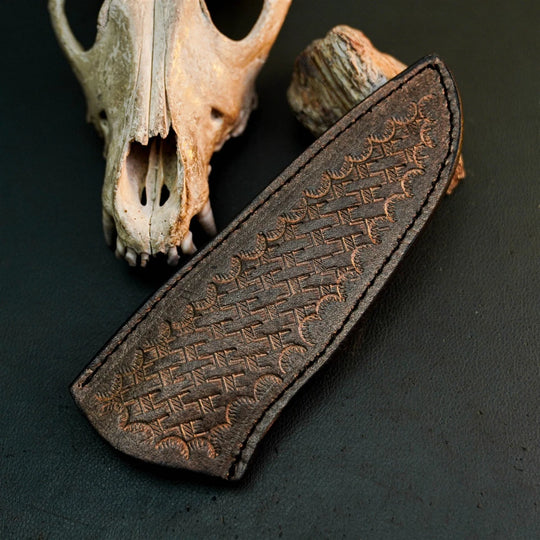 A dark brown leather knife sheath with an embossed geometric pattern, displayed diagonally on a dark textured surface with a weathered animal skull and wood prop in the background.