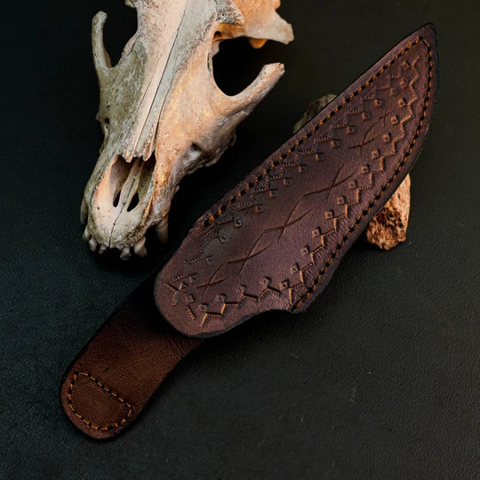 A dark brown leather knife sheath with embossed geometric patterns and contrast stitching, displayed diagonally on a dark textured surface next to a weathered animal skull.