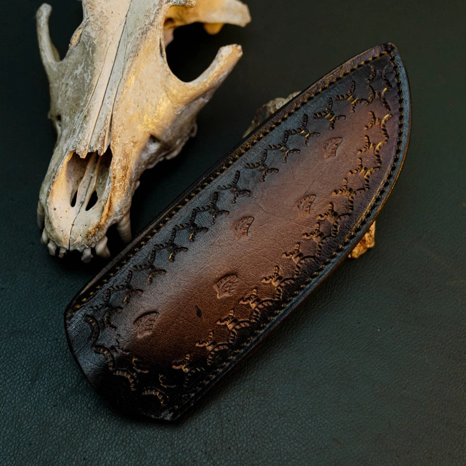 A dark brown leather knife sheath with embossed floral patterns, displayed diagonally next to a weathered animal skull on a dark textured background.