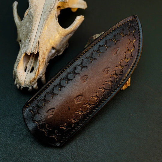 A dark brown leather knife sheath with embossed floral patterns, displayed diagonally next to a weathered animal skull on a dark textured background.