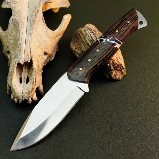 A fixed-blade knife with a polished silver blade and a dark brown wood handle with brass pins, displayed diagonally on a dark textured surface next to a bone skull and wood prop.