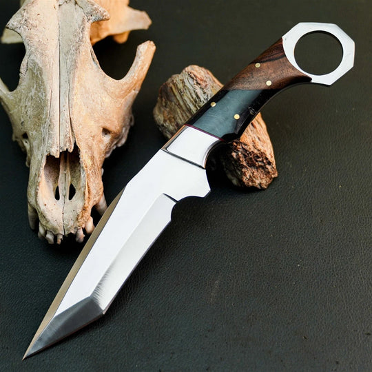 A fixed-blade knife with a polished silver blade and a dark wood and black handle, styled on a dark textured surface next to a bone skull and wood props.