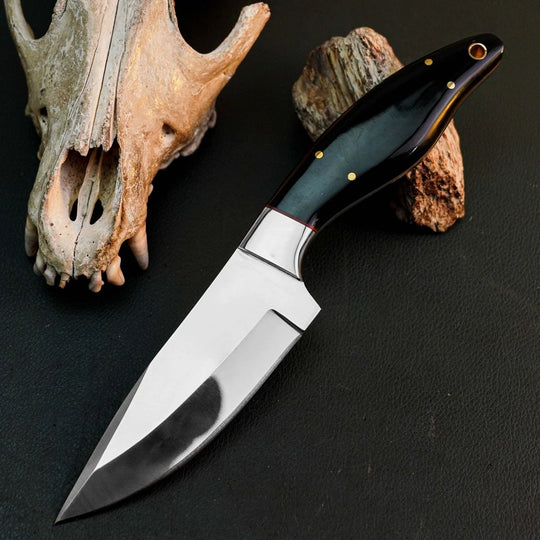 A fixed-blade knife with a polished silver blade and a dark black handle with brass pins, displayed diagonally on a dark textured surface next to a deer skull and a piece of wood.