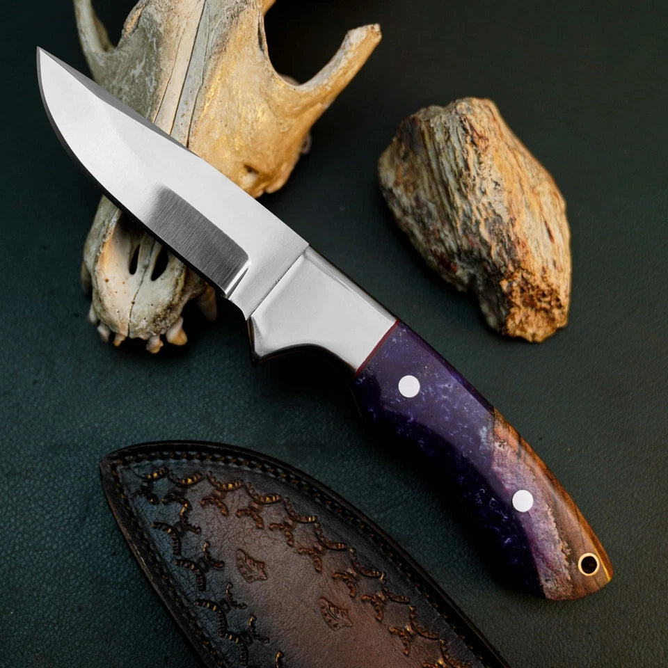 A fixed-blade knife with a satin-finished silver blade and a purple marbled handle, displayed diagonally on a dark textured surface with a leather sheath and natural animal bones as props.