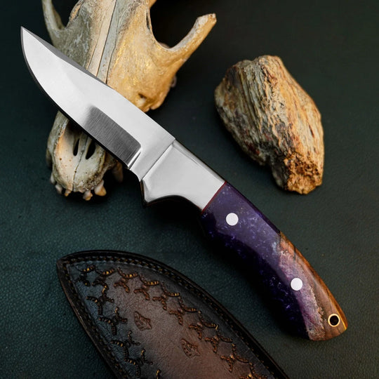 A fixed-blade knife with a satin-finished silver blade and a purple marbled handle, displayed diagonally on a dark textured surface with a leather sheath and natural animal bones as props.