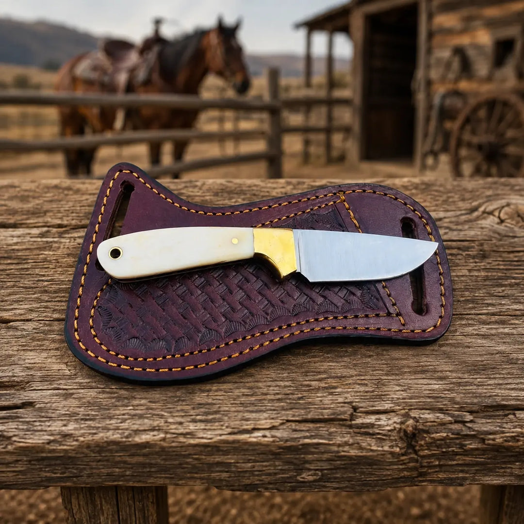A fixed-blade knife with an ivory-colored handle and gold bolster rests on a dark brown leather sheath, placed on a rustic wooden table with a blurred western ranch background featuring a horse and a wagon wheel.