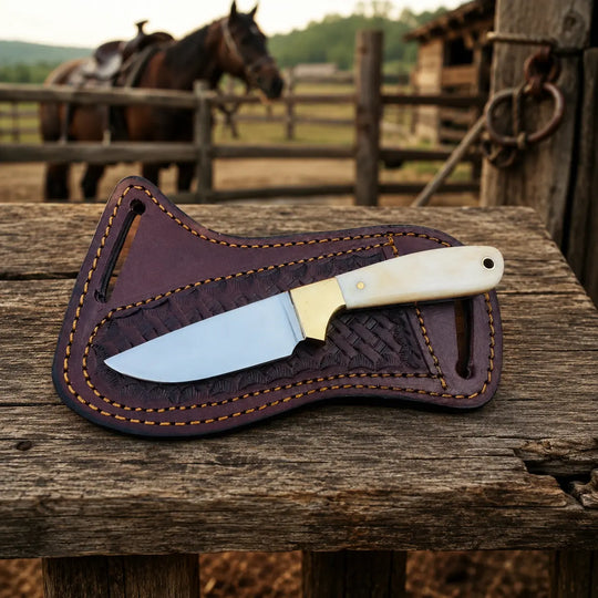 A fixed-blade knife with a silver blade and cream handle rests on a dark brown leather sheath with tan stitching, placed on a rustic wooden fence in an outdoor barn setting with horses in the background.