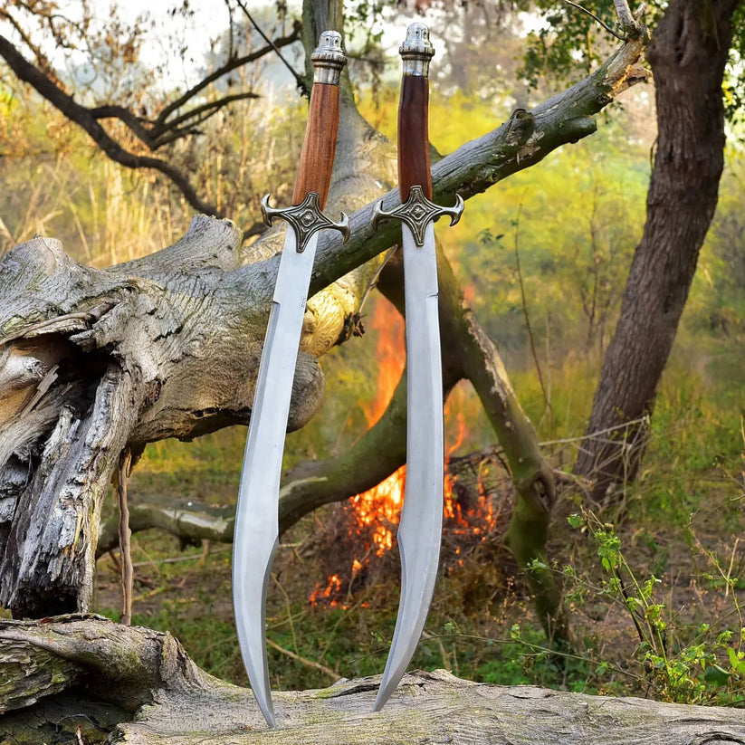 Two curved steel daggers with wooden handles and ornate silver hilts are displayed upright on a fallen log in an outdoor forest setting with a fire burning in the background.
