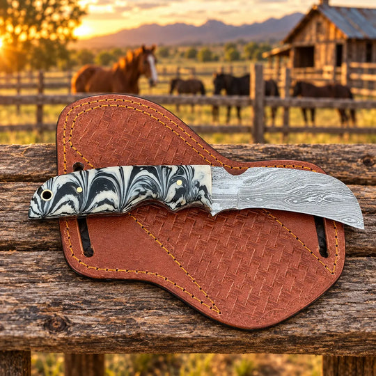 A fixed-blade knife with a black, white, and gray marbled handle and a patterned steel blade rests on a brown leather sheath placed on a rustic wooden fence. The background shows a blurred outdoor ranch scene at sunset with horses and a barn.
